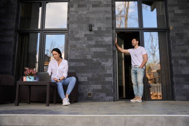 Adam, pencerenin yanında otururken dizüstü bilgisayarda yazan kadına bakıyor.