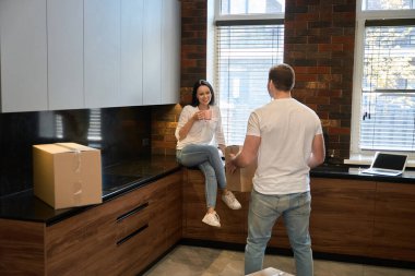 Elinde kupa olan esmer kadın mutfaktaki komodinin üzerinde oturuyor.