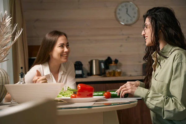 微笑着的女性朋友们坐在桌旁准备食物 互相望着对方 — 图库照片