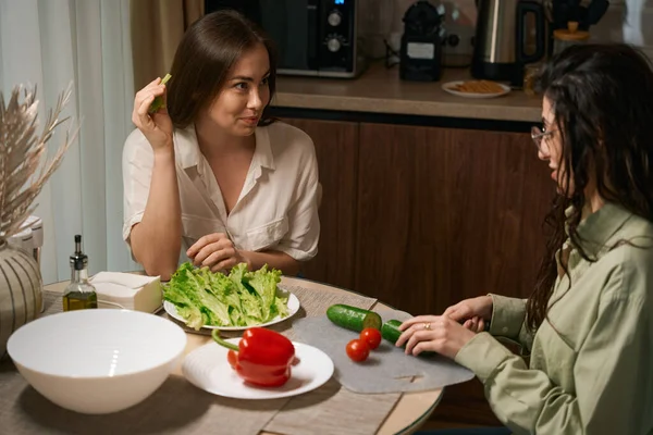 台所の女性と話をしながらテーブルに座って食事を準備している眼鏡の女性 — ストック写真