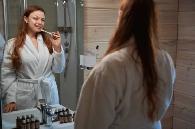 Elinde diş fırçası olan odaklanmış bir kadın banyo aynasında kendine bakarken lavaboya yaslanıyor.