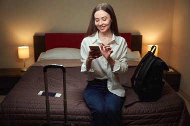 Yanında bavul ve sırt çantasıyla kanepede oturan mutlu bayan telefona göz atıyor. Bir otel odasına giriş yap.
