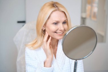 Mutlu olgun kadın aynaya bakıyor ve tek eliyle yüzüne dokunuyor.