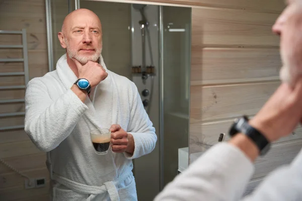 手里拿着咖啡杯 凝视着浴室镜子里自己的脸的男人 — 图库照片