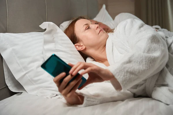 认真的女性 手拿手机躺在床上看着熟睡的配偶 — 图库照片