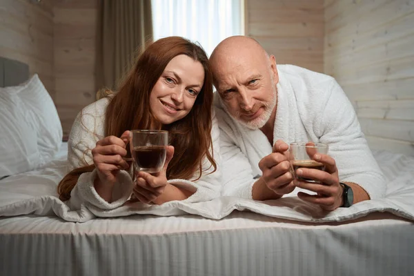Ευτυχισμένοι Σύζυγοι Μπουρνούζια Ξαπλωμένοι Στο Κρεβάτι Φλιτζάνια Καφέ Στα Χέρια — Φωτογραφία Αρχείου