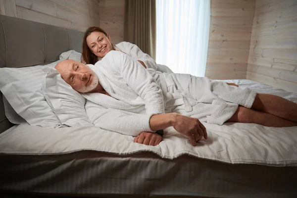 一位微笑的女士一边躺在丈夫身边一边拍拍丈夫的肩膀 一边躺在床上 — 图库照片