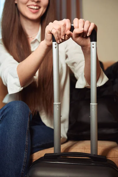 stock image Smiling young woman holding the handle of a luggage sitting on a sofa in a hotel room. Cropped photo