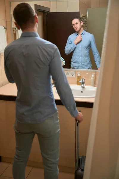年轻的小伙子站在浴室的洗脸盆边 镜中看着自己 提着手提箱 入住酒店房间 — 图库照片