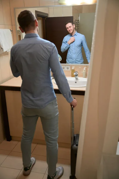 一个英俊的男人站在浴室的洗脸盆边 站在镜子里 提着行李 入住酒店房间 — 图库照片