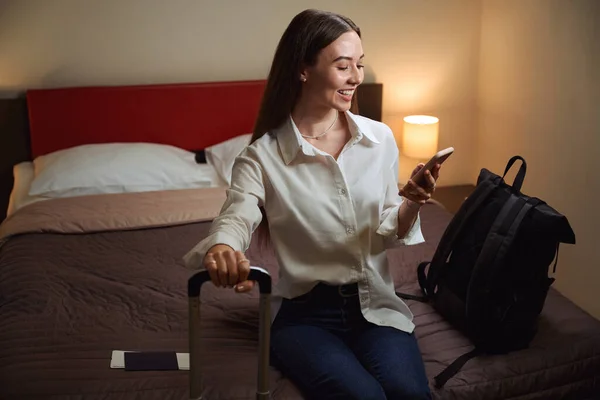 ホテルの部屋でソファに座っている間 幸せな女性は電話をブラウジングし スーツケースハンドルを保持 — ストック写真