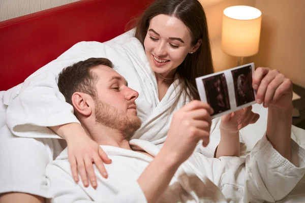 微笑的年轻女人躺在酒店房间的床上 一边用超声波照片指着胎儿 一边对着她的伴侣 — 图库照片