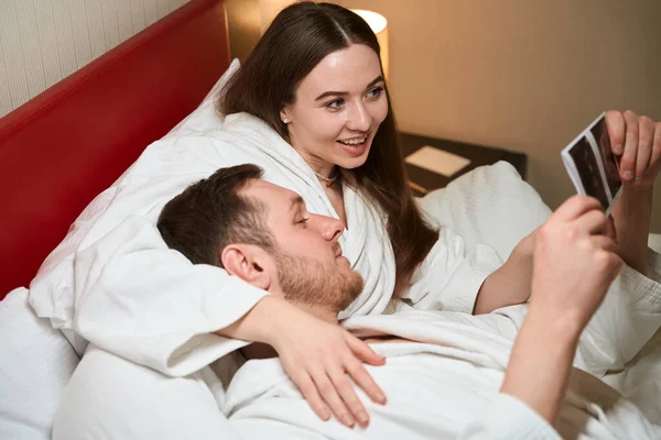 快乐的女性在酒店房间向躺在床上的伴侣展示未来宝宝的超声照片 — 图库照片