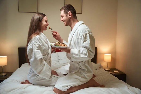 快乐的年轻人在宾馆的套间里给他快乐的妻子吃美味的煎饼 — 图库照片