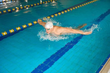 Beyaz şapkalı yetişkin sporcu su egzersizleriyle meşgul, kelebek stilinde yüzüyor.