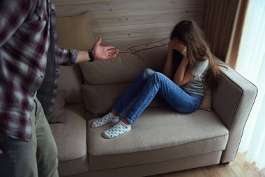 Kanepede oturan, elleriyle yüzünü örten ve ailesinin önünde ağlayan bir kız.