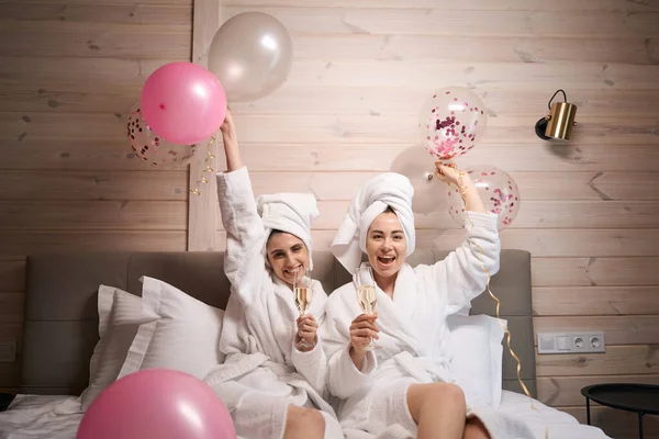 Dvě Usměvavé Dámy Balónky Sklenicemi Šampaňského Sedí Velké Posteli Hotelovém — Stock fotografie