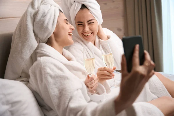 两个穿着白色浴衣的漂亮女人躺在豪华酒店房间里 并在鸡舍派对上用智能手机拍照 — 图库照片