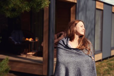 Kareli kumaşa sarılı sakin bir kadın, ahşap kulübenin yanında dururken yüzünü güneşe çeviriyor.