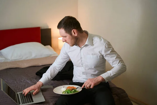 Serious young man seated on bed in hotel room pricking food with fork and typing on laptop keyboard