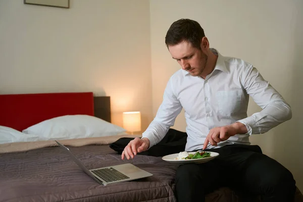 男子坐在一台便携式计算机前的床上 用叉子挑食 — 图库照片
