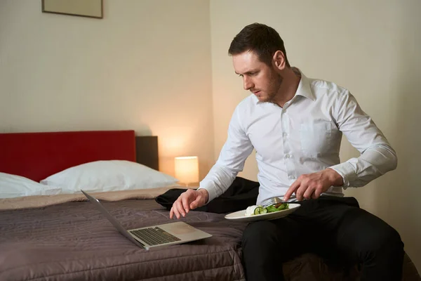 商人坐在旅馆房间的床上 一边用叉子叉着食物 一边看笔记本电脑屏幕 — 图库照片