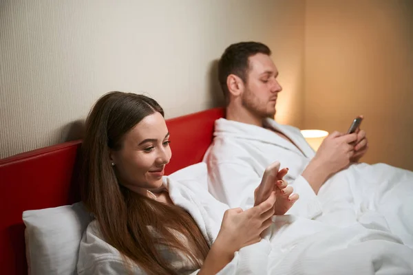 穿着浴衣的年轻夫妇躺在酒店房间的床上 手里拿着手机 — 图库照片