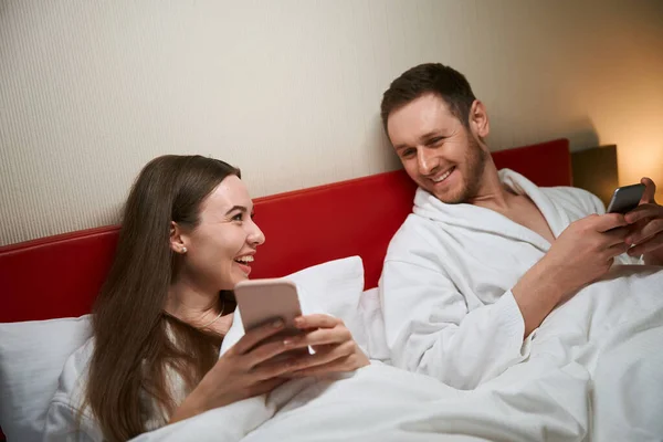 快乐的年轻女性在智能手机屏幕上向躺在床上的快乐伴侣展示一些东西 — 图库照片