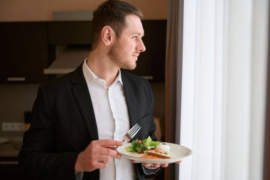 Takım elbiseli bir adam elinde yemek tabağı ve otelin penceresinden dışarı bakıyor.