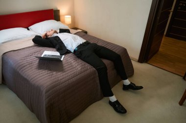 Takım elbiseli genç bir erkek otel odasında dizüstü bilgisayarın yanında yatıyor.
