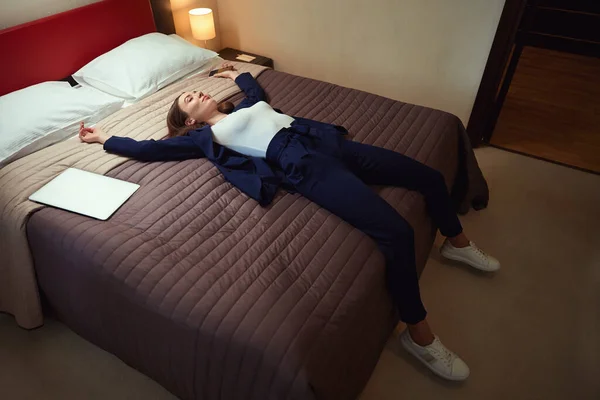 彼女の手の携帯電話とのビジネスススーツの若い女性は モーテルのラップトップの近くのベッドにあります — ストック写真