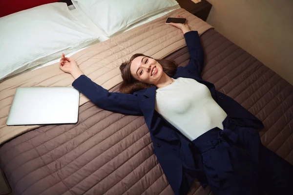 モーテルでベッドで休んでいる彼女の手のスマートフォンとのビジネスススーツの女性 カメラを見て — ストック写真