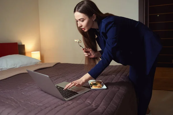 ベッドの近くに立って食べるビジネスススーツの女性 ホテルの部屋でコンピュータで働く — ストック写真
