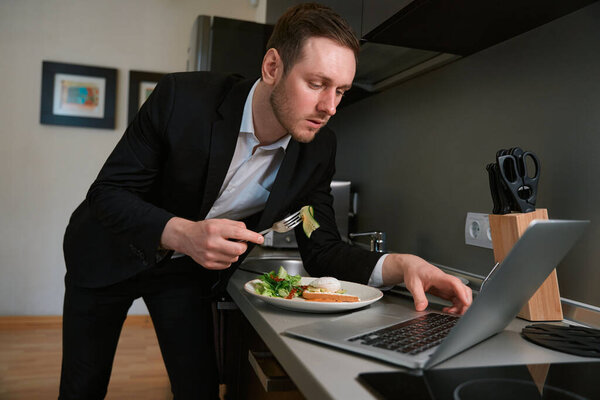 Young male in business suit standing near table and eating, typing on laptop in the hotel room