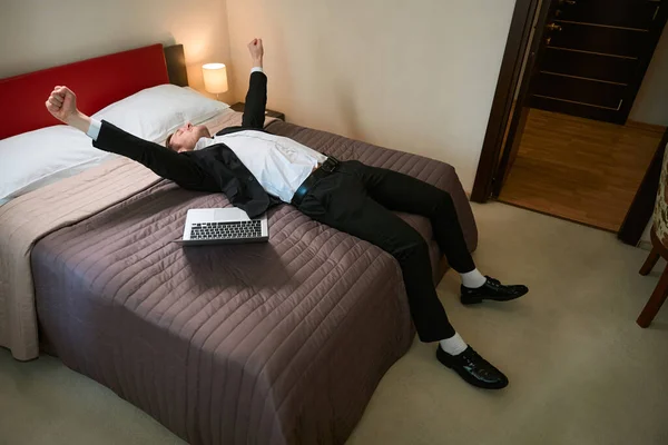 ビジネスススーツの若い男性は ラップトップの近くのベッドに横たわり モーテルルームで休む喜び — ストック写真