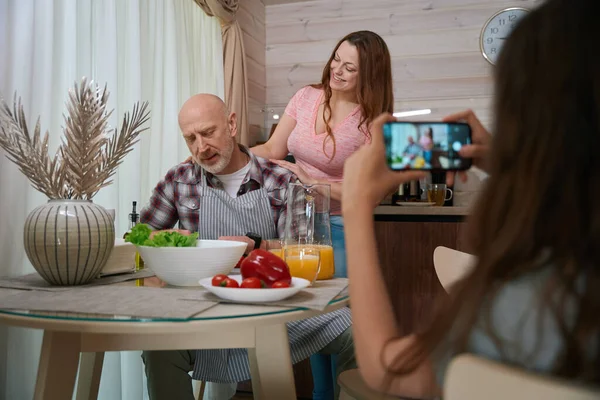 stock image Girl photographing with smartphone her smiling mother standing beside her busy father preparing vegetable salad