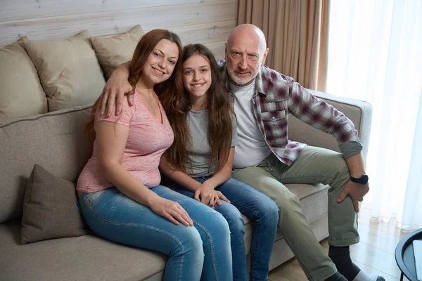Calm mature man seated on comfortable sofa hugging his smiling wife and cheerful daughter