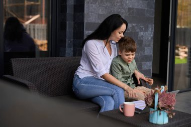 Evin önündeki masada oturan bir çocuk ve genç bir kadın dizüstü bilgisayara bakıyorlar.