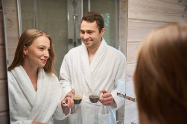 Diş fırçalı bir kadın ve banyoda aynanın yanında kahve standı olan bir adam.