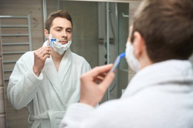 Yüzünde köpük olan genç adam aynaya bakarak sakalını tıraş ediyor.