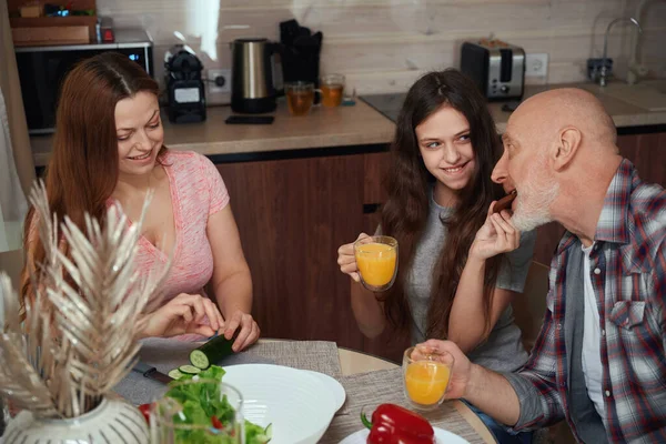 stock image Loving daughter feeding her father with oatmeal cookie at table while her mother making salad