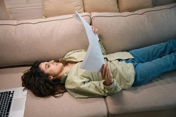 年轻的女士躺在沙发上 手里拿着报告 正在用笔记本电脑远程办公 — 图库照片