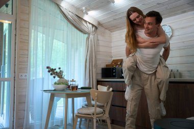 Genç bir kadın mutfaktaki bir adamın sırtına biniyor, çift birbirine bakıyor.