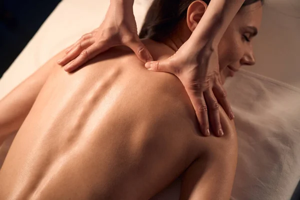 Masseuse hands pressing into trapezius muscle knot on female patient upper back