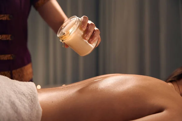 Cropped photo of masseuse dripping melted wax from jar on naked back of woman