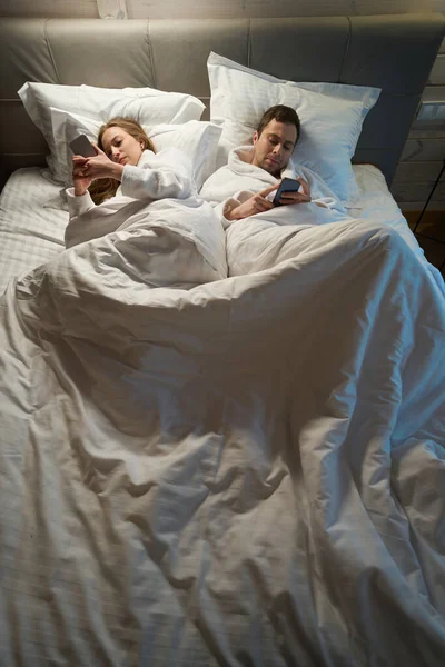 Indifferent spouses lying apart in big comfortable bed and playing on smartphones, lack of common interests, misunderstanding in relationships