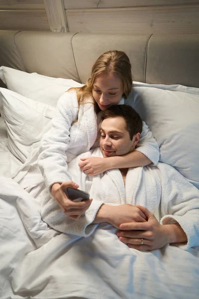 Happy young couple in love making selfie on smartphone, wife in soft bathrobe hugging her lovely husband and posing, spouses having fun spending time together