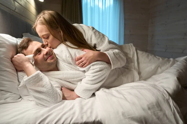 Sensual attractive woman tenderly kissing her husband in cheek, suggesting to cuddle, newlywed couple lying in bed in soft bathrobes, romance, love and passion