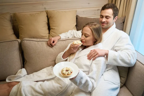 英俊的男人笑着看着他年轻的妻子靠着他吃甜蛋糕 新婚夫妇穿着舒适的浴衣坐在沙发上休息 — 图库照片