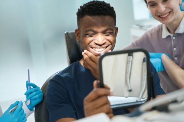 Stomatologlardan oluşan bir ekip, Afro-Amerikan erkek müşterilerinin işlerinden memnun olmalarını izliyor. Aynadaki dişlere bakıp gülümsüyor.